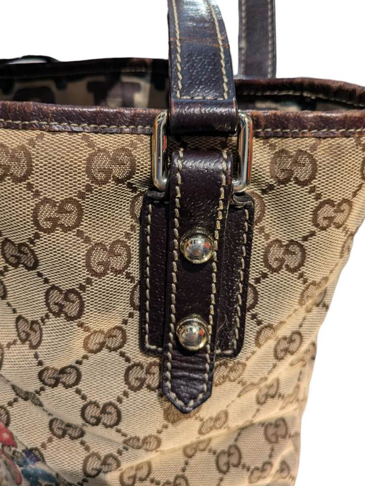 Gucci GG Canvas Mushrooms tote bag brown canvas and brown leather bag with an embroidered red mushroom design and a mushroom charm, white, purple, and green pattern fabric interior. Close up of buckle with gold hardware