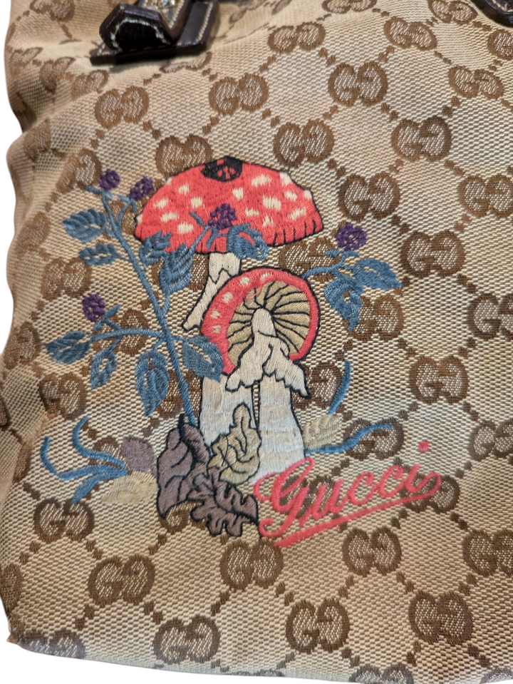 Gucci GG Canvas Mushrooms tote bag brown canvas and brown leather bag with an embroidered red mushroom design and a mushroom charm. Close up of embroidery with red and white mushrooms with speckled tops, brown leaves, and green leaves and " Gucci" in red