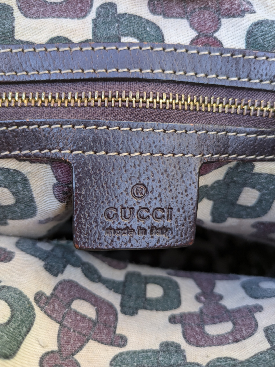 Gucci GG Canvas Mushrooms tote bag brown canvas and brown leather bag with an embroidered red mushroom design and a mushroom charm, white, purple, and green pattern fabric interior 
Gucci label